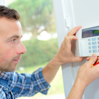 A man wearing a blue plaid shirt installing a burglar alarm on an interior wall near an open doorway.