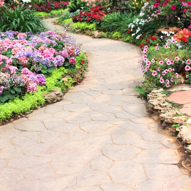 A tan stone walkway stretching across a residential garden. Colorful flowers line both sides of the walkway.