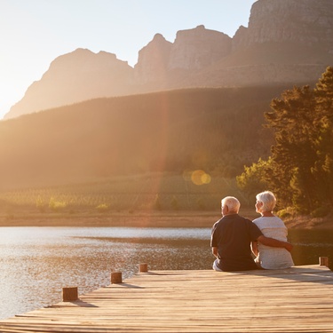 A senior couple sits on a wooden dock overlooking a calm lake and mountains as the sun sets in the distance.