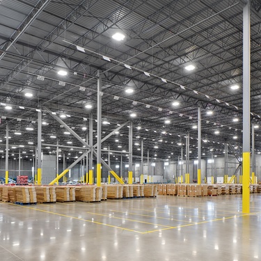 A brightly lit warehouse with shiny, reflective floors, forklifts, and stacks of boxes and plywood in the middle of it.