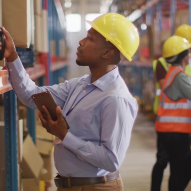 A warehouse supervisor in a yellow hard hat scans items with a barcode scanner and tablet as employees work behind him.