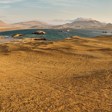 A traveler stands at the base of a sandy hill, overlooking a coastline in the distance as other travelers approach it.