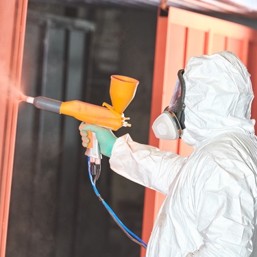 A person wearing full PPE applies an orange protective finish to a sheet of metal using a powder coating gun.
