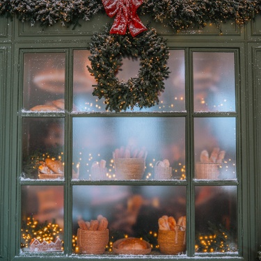A green wreath with a red ribbon hangs on a frosted window, displaying lit baskets of baked goods behind the glass.