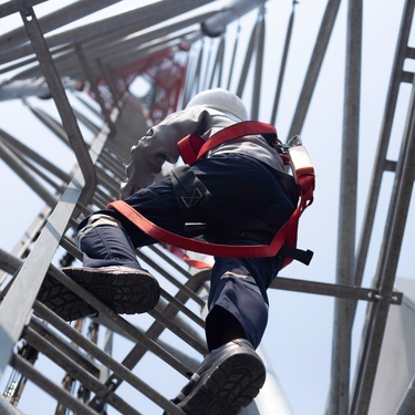 An engineer climbing up the center ladder of a communication tower. They're using a red safety harness to attach themselves.