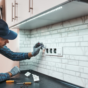 A handyman wears a baseball cap as he leans under the cabinets to adjust the electrical boxes on the backsplash.