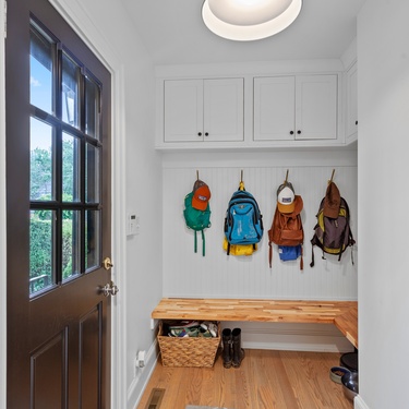 A nook in the corner of the front door area, serving as a mudroom with four backpacks hanging on hooks and a bench seat.