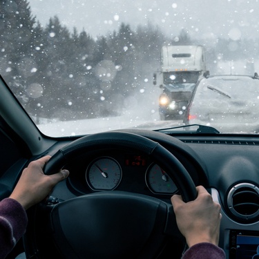 A driver holds the steering wheel with both hands as they travel in a snowstorm. There is an SUV in front of them.