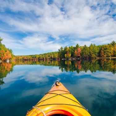A yellow kayak sits on a large body of water that's surrounded by trees. The trees contain fall-colored leaves.