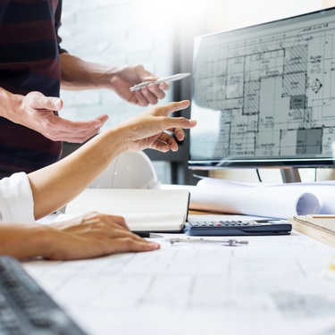 A pair of construction engineers at the desk, reviewing blueprints and design plans on the computer monitor.