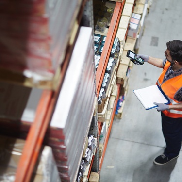 A worker in a warehouse is in an orange high-visibility vest and scans different boxes on some tall shelves.