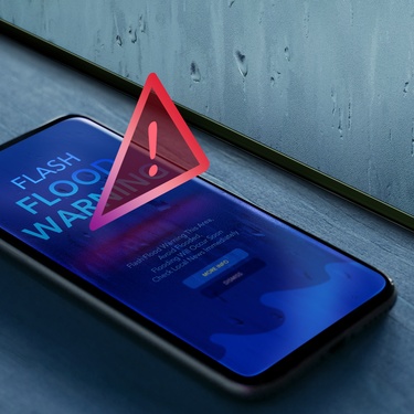 A smartphone laying on a windowsill near a wet window displays a flash flood warning with a red triangle alert icon.