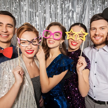 A group of five party guests smiling and posing in a photo booth. Each guest is holding a small handheld prop.