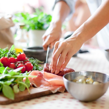 A homeowner chopping ingredients with a knife in their kitchen. The ingredients include tomatoes, salami, and more.
