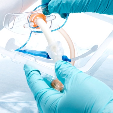 Someone wearing latex gloves holds tracheostomy equipment in a package. The package is open, and they lift the tube out.