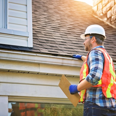 A man in a vest and hard hat stands beside the roof of a home. He is resting his hand on the gutter.