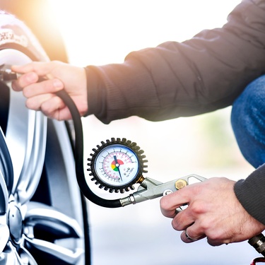 A car owner kneeling down next to their car's tire. They're using a tire pressure gauge to measure the psi.