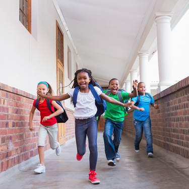 Four schoolchildren wearing backpacks run down a covered brick hallway, smiling and laughing together.