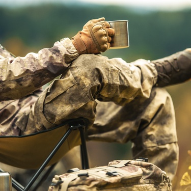 A solo hunter sitting on a camping chair and looking out to the wilderness while drinking from a metal cup.