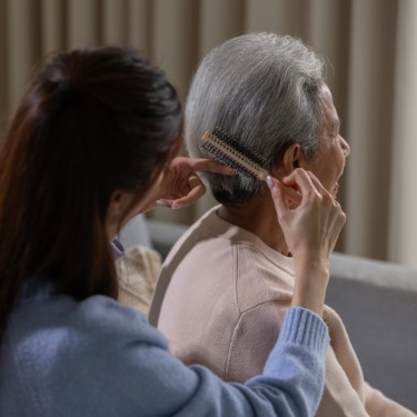 An aging mother sits quietly as her younger daughter combs her hair with a brush. She has short, gray hair.