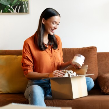 Woman smiling while opening a shipping box on a sofa, holding a round jar with tissue paper inside the package.