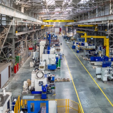A large metal manufacturing floor with overhead cranes, blue machinery, yellow columns, work areas, and marked lanes.