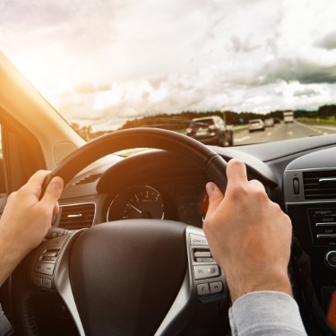 Driver's hands grip the steering wheel while traveling on a highway beneath dramatic clouds with several vehicles ahead.