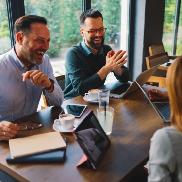 Four adults in a casual business meeting laughing around a cafe table with laptops and coffee cups in front of them.