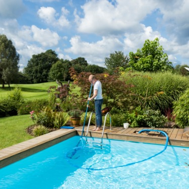 A person cleaning a backyard swimming pool with a long vacuum and attached hose while surrounded by lush garden landscaping.