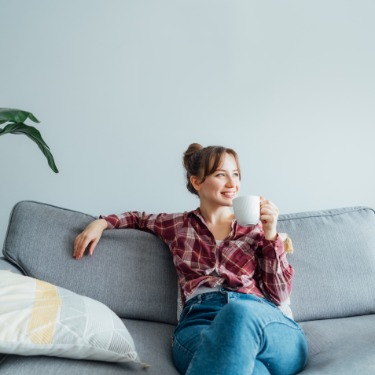 A smiling brunette woman in a red plaid shirt and blue jeans sitting on a gray sofa, holding a white mug beside cushions.