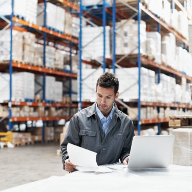 A man in a gray work jacket reviewing papers beside an open laptop in a warehouse lined with tall shelves of boxes.