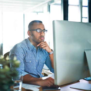 Man wearing glasses sits at an office desk, focusing on a large desktop computer monitor in a bright workspace.