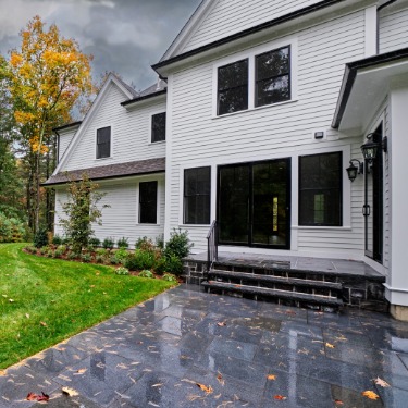A wet stone patio behind a white house, with black-framed windows, green grass, and autumn trees beneath gray clouds.