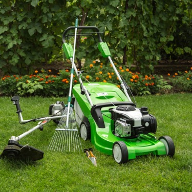 A green lawn mower, string trimmer, rake, and hand tools arranged together on grass in a landscaped garden.