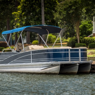 Blue pontoon boat with canopy tied to a wooden dock on a calm lake, surrounded by trees and lakeside homes.
