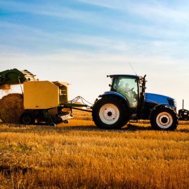 A side view of a blue tractor towing a yellow round hay baler as it drops a round hay bale in an empty field.