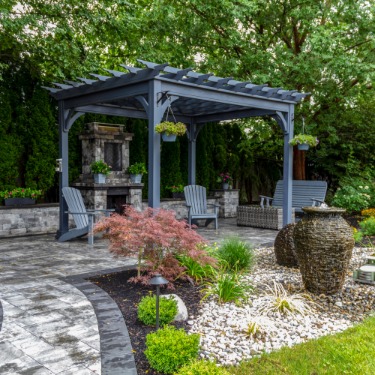 A backyard with lush green grass, a tiled pathway leading to a wooden pergola over multiple chairs and a fireplace.