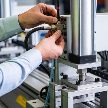 A person wearing a high-visibility vest adjusting a hose fitting on a pneumatic cylinder beside metal machinery.