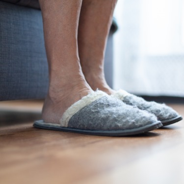 A close-up of two feet wearing fluffy slippers on a vinyl brown floor next to a bed with a grey boxspring.