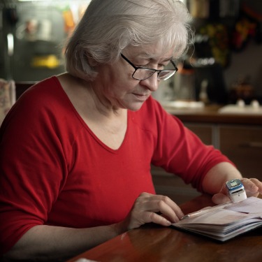A senior woman sits at a table with a pulse oximeter on the index finger of her left hand. A notebook is open on the table.