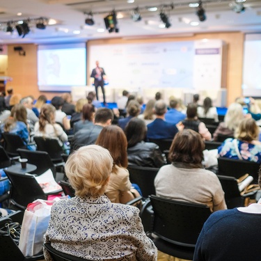 A room full of people sitting and looking at a presenter at the front, standing on a platform with a white background.