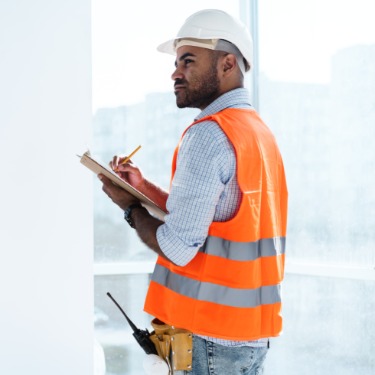 A man standing near a window with a high-visibility vest, white hard hat, and utility belt writes on a clipboard.