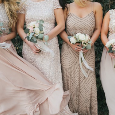 Four women in light tan dresses standing together and holding small bouquets of white and pink flowers.