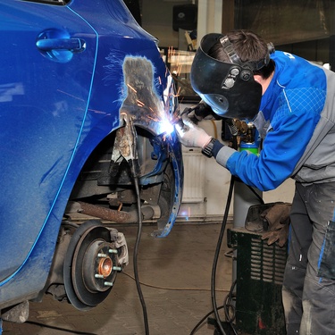 A person wearing a welding mask and gloves is welding a piece of metal on the wheel well of an elevated blue car.