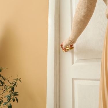 A person in a beige sweater and pleated skirt holds the gold handle of a white interior door. A plant is near the wall.