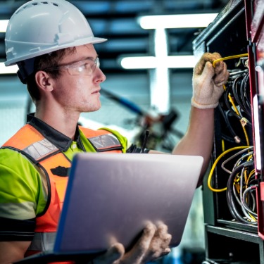 A man wearing a white helmet, holding a laptop while checking the connections inside a small electrical panel box.