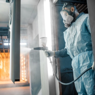 A technician in protective gear sprays a coating onto a metal part inside an industrial spray booth with bright work lights.