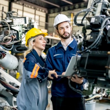 A man and a woman are standing in an automotive facility. They’re looking at an engine suspended by a chain.