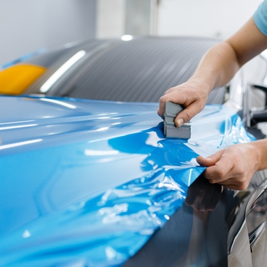 A person uses a squeegee to install a bright blue vinyl wrap onto the hood of a black vehicle in an auto body shop.