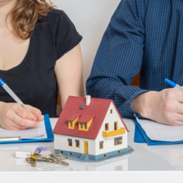 A man sitting next to a woman behind a desk while both hold pens over documents with a small house in the middle.